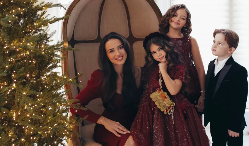 A mum, her two daughters and her son pose next to an indoor Christmas tree decorated with fairy lights. The girls are all dressed in burgundy dresses and the boy is in a smart black suit and white shirt.
